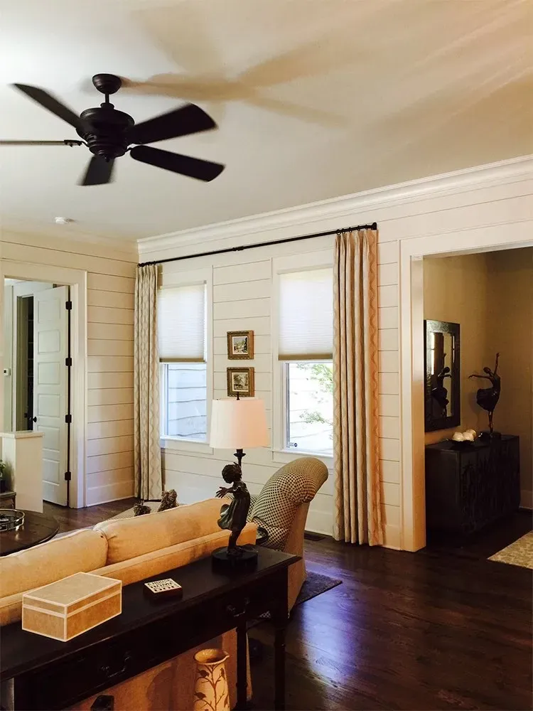 A cozy living room with white shiplap walls, dark hardwood floors, a ceiling fan, and beige curtains framing two windows.
