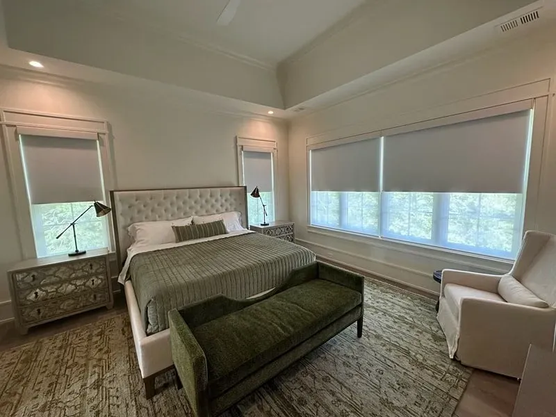 A bedroom with a tufted headboard, olive-green bed runner, matching bench, bedside lamp, and a white armchair by windows.