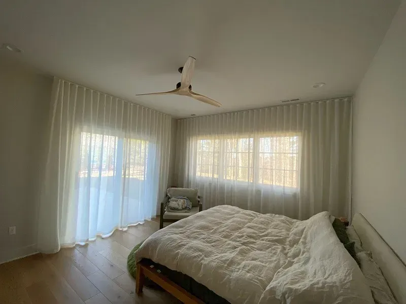 A sunlit bedroom features a bed with white bedding, sheer curtains covering large windows, and a ceiling fan overhead.
