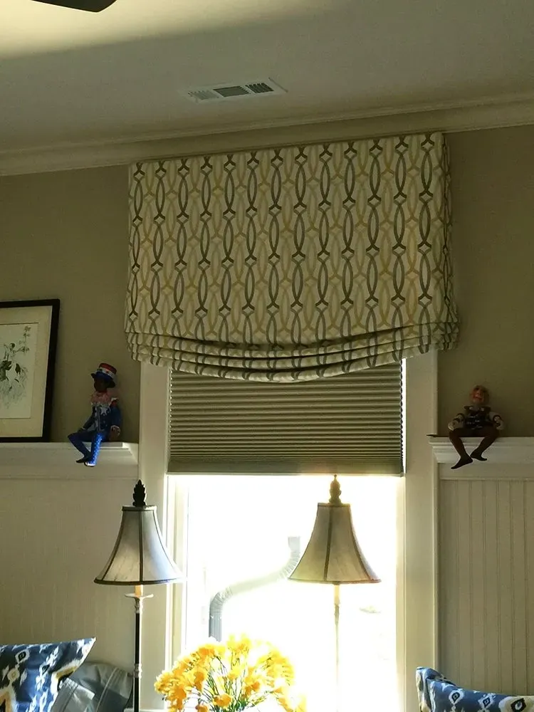 Window with layered patterned valances, a blind, lamps, and small figurines on shelves in a living room.