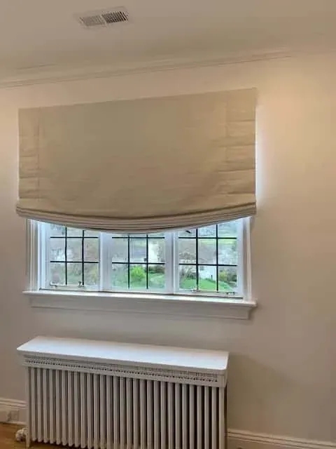 A white wall with a window covered by a partially raised, light beige Roman shade above a white radiator cover.