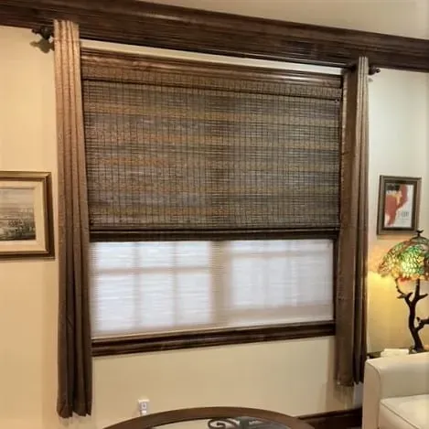 A window with a woven wood shade layered over a white cellular shade, flanked by brown curtains under a wooden valance.