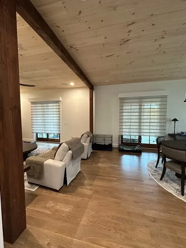 A sunlit living room with a white armchair, light wood plank ceiling, exposed dark wood beams, and a pet crate.