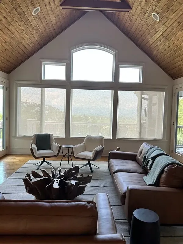 A sunlit living room with a brown leather sofa, two white chairs, and a large wooden coffee table under a vaulted ceiling.