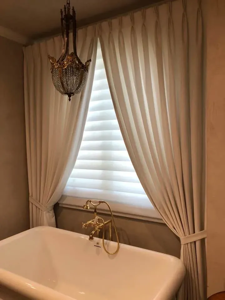A white bathtub with gold fixtures in front of a window with white shutters and floor-length cream curtains.