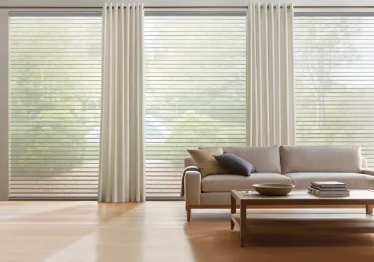 A bright living room with horizontal window blinds, light curtains, a sofa with pillows, and a wooden coffee table.