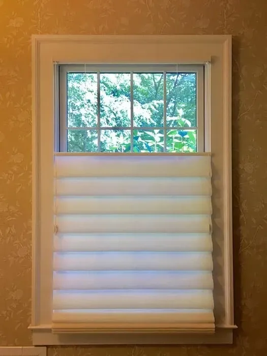 A window with a white, bottom-up cellular shade covering the lower half, framed by light gold patterned wallpaper.