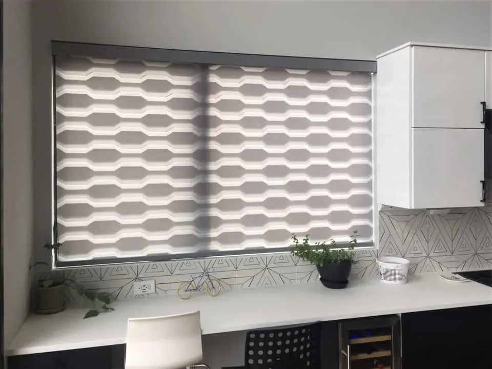 A light-colored desk in a modern kitchen with a window featuring gray and white geometric patterned roller shades.