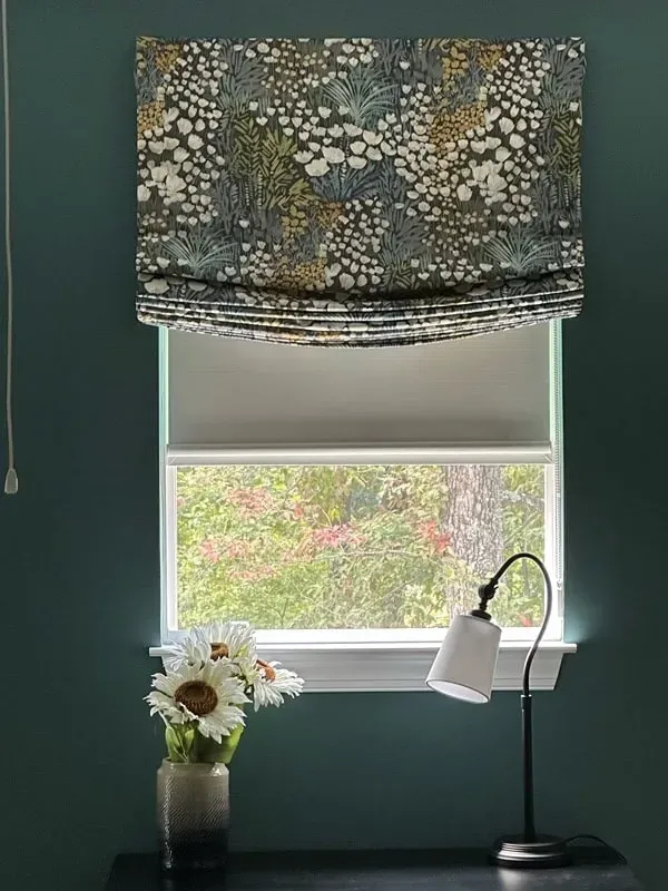 A patterned Roman shade hangs over a window with a view of greenery, beside a lamp and a vase of flowers on a table.