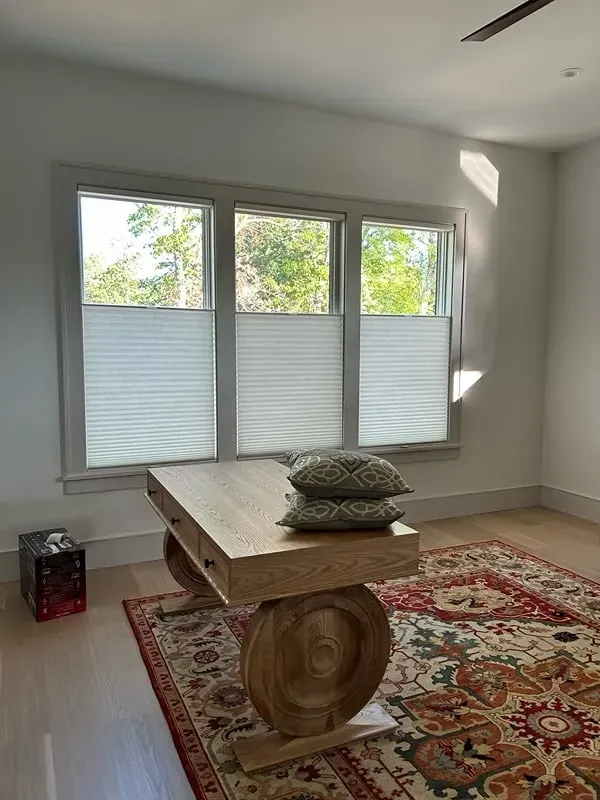 A wooden desk with a circular leg base sits on a colorful patterned rug in a sunlit room with window shades.