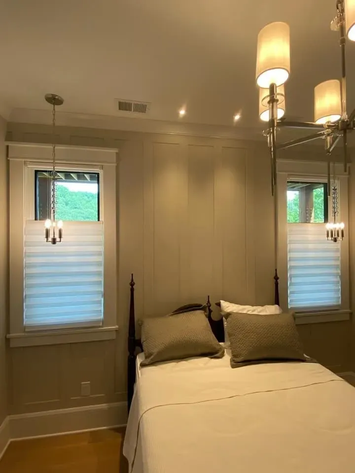 A bedroom featuring two windows flanking a bed, adorned with light-colored bedding, neutral walls, and hanging chandeliers.