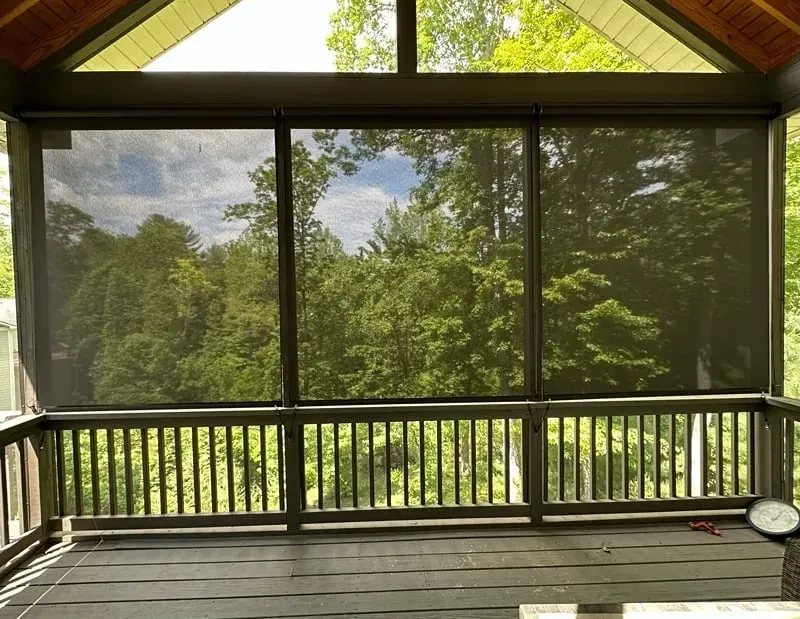A view from a wooden porch through dark solar shades, looking out at lush green trees under a bright sky.