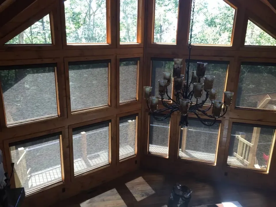 A view from inside a room through a large, multi-paned wooden window frame, showing trees and a deck with a chandelier.