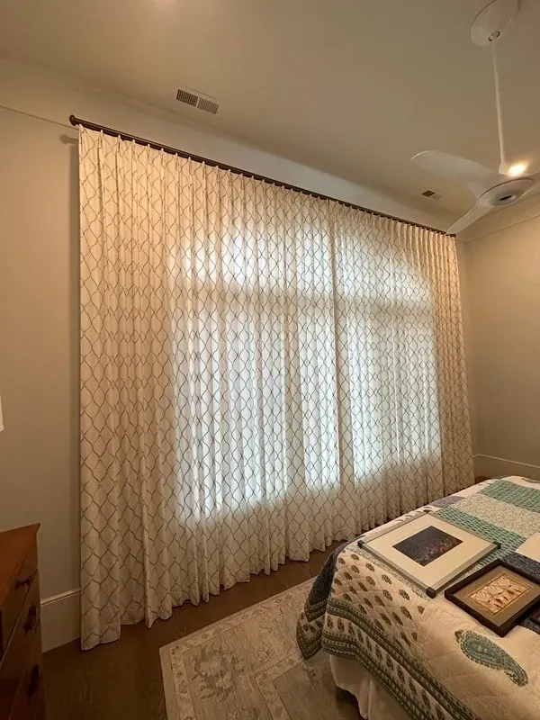 Floor-to-ceiling patterned curtains hang on a rod against a light wall next to a bed in a bedroom.
