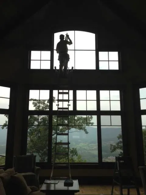 A person stands on a tall ladder inside a home, cleaning the large arched window overlooking a mountain landscape.