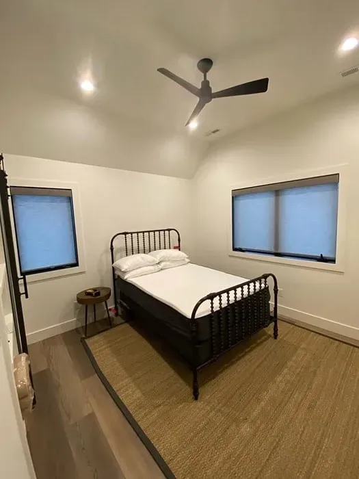 A minimalist bedroom with a black metal bed, white bedding, a natural fiber rug, and two windows under a vaulted ceiling.