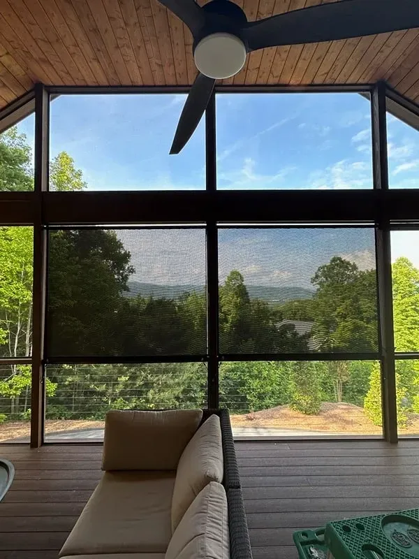 A wooden deck with a couch, viewed through large windows with solar shades overlooking a mountain forest landscape.