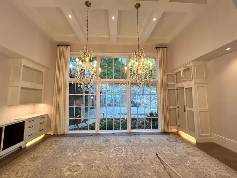 A bright room with a large window, coffered ceiling, two crystal chandeliers, patterned rug, and white built-in cabinets.