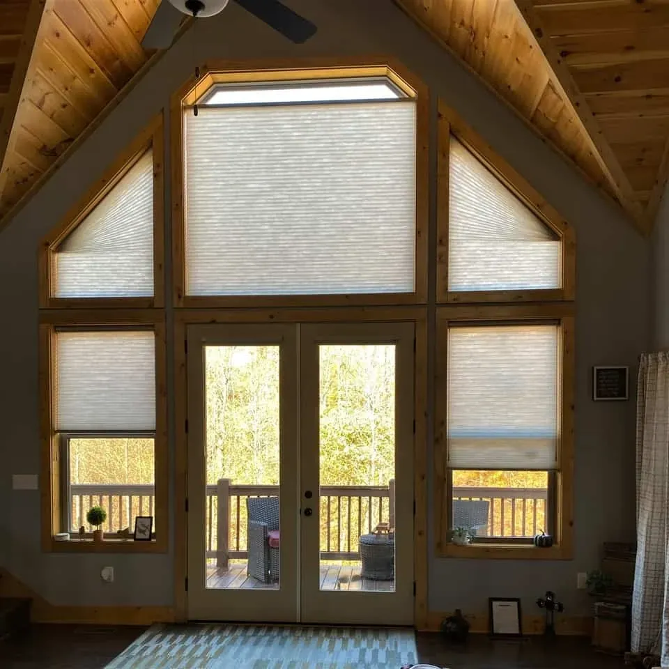 Large windows with closed beige cellular shades, surrounding a glass door in a room with a vaulted wood-paneled ceiling.