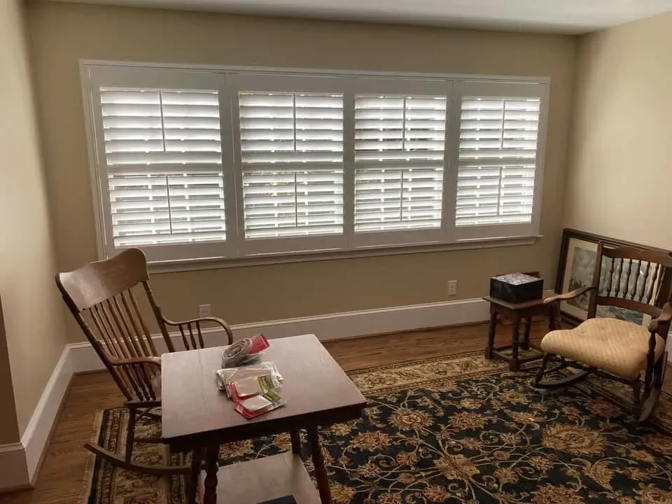 Two wooden rocking chairs facing each other in a room with a large, white-shuttered window and a patterned rug.