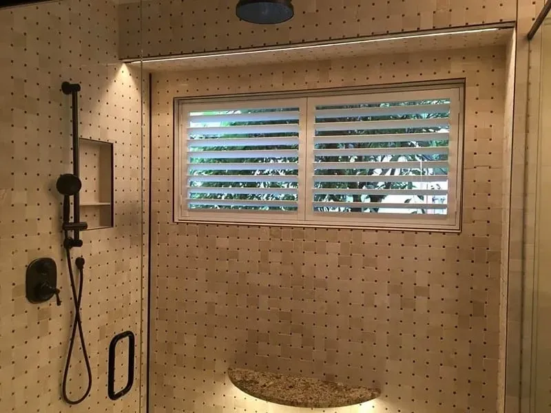 A tiled shower with a black handheld showerhead, a recessed shelf, a wall-mounted seat, and a window with white shutters.
