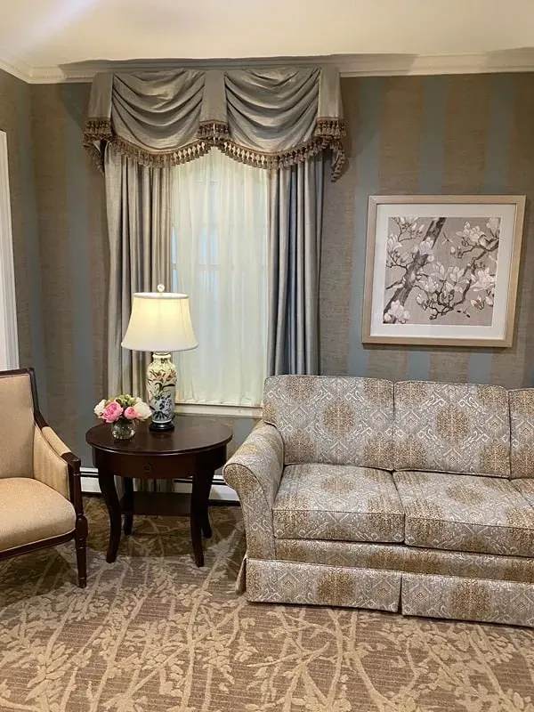 A seating area with a patterned sofa, a side table with a lamp and flowers, a chair, and window curtains in a gray room.