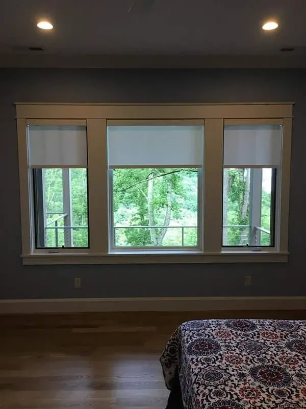 A room features a large, three-section window with white trim, white roller shades, and a view of trees, plus a patterned bed.