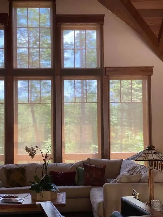 A cozy living room with a large sectional couch and high, wood-framed windows looking out onto a sunny, wooded area.