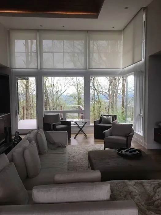 Modern living room with a large sofa, ottoman, and armchairs facing a wall of windows overlooking a wooded landscape.