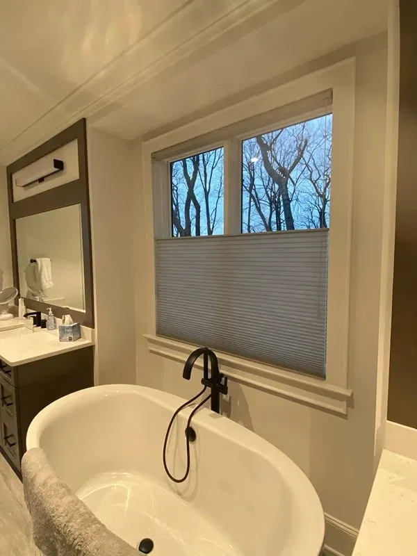 A bathroom interior featuring a white freestanding bathtub with a black faucet, set below a window with a pleated shade.