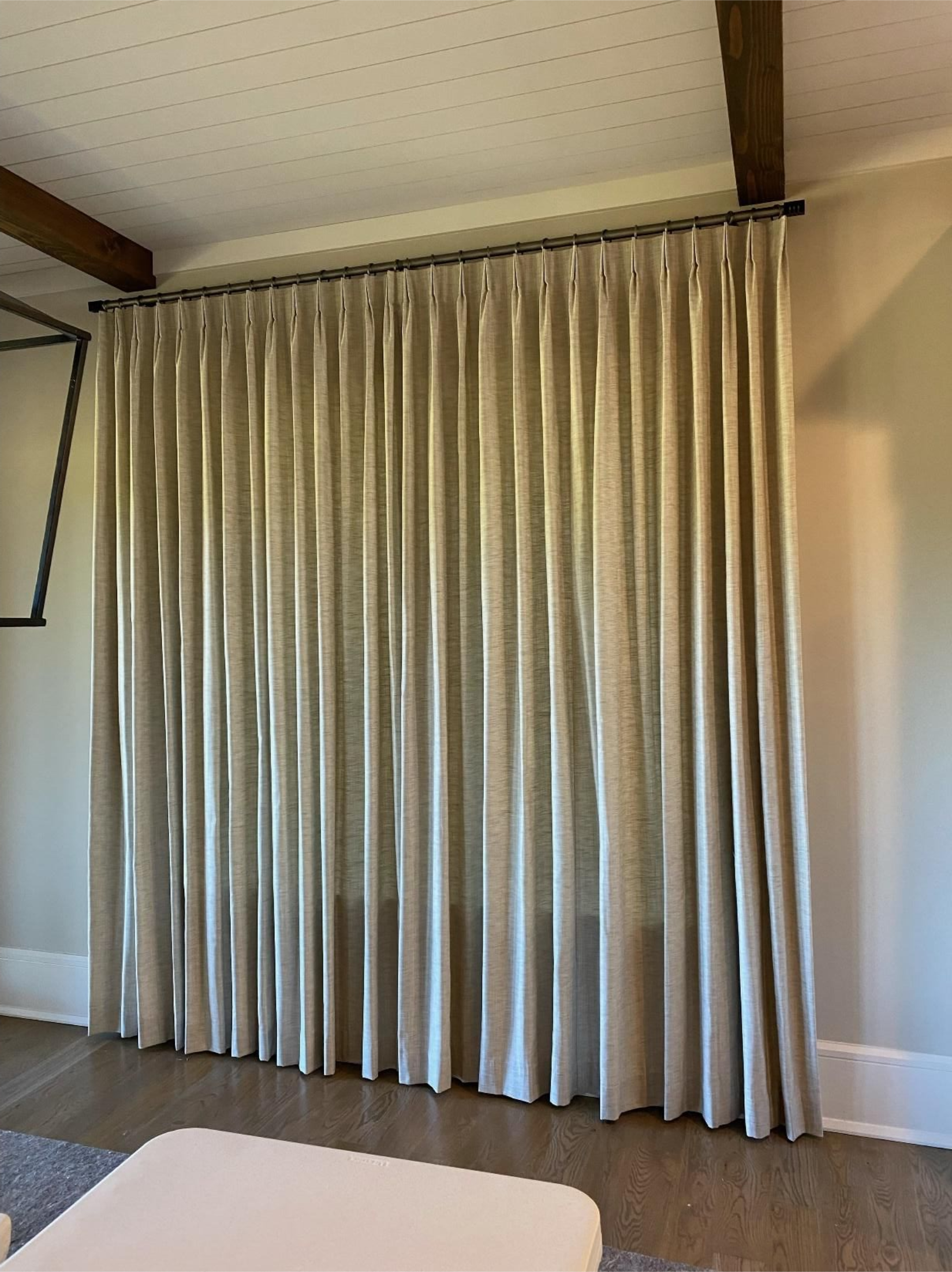 Beige curtains covering a large window in a bright room with wooden ceiling beams.