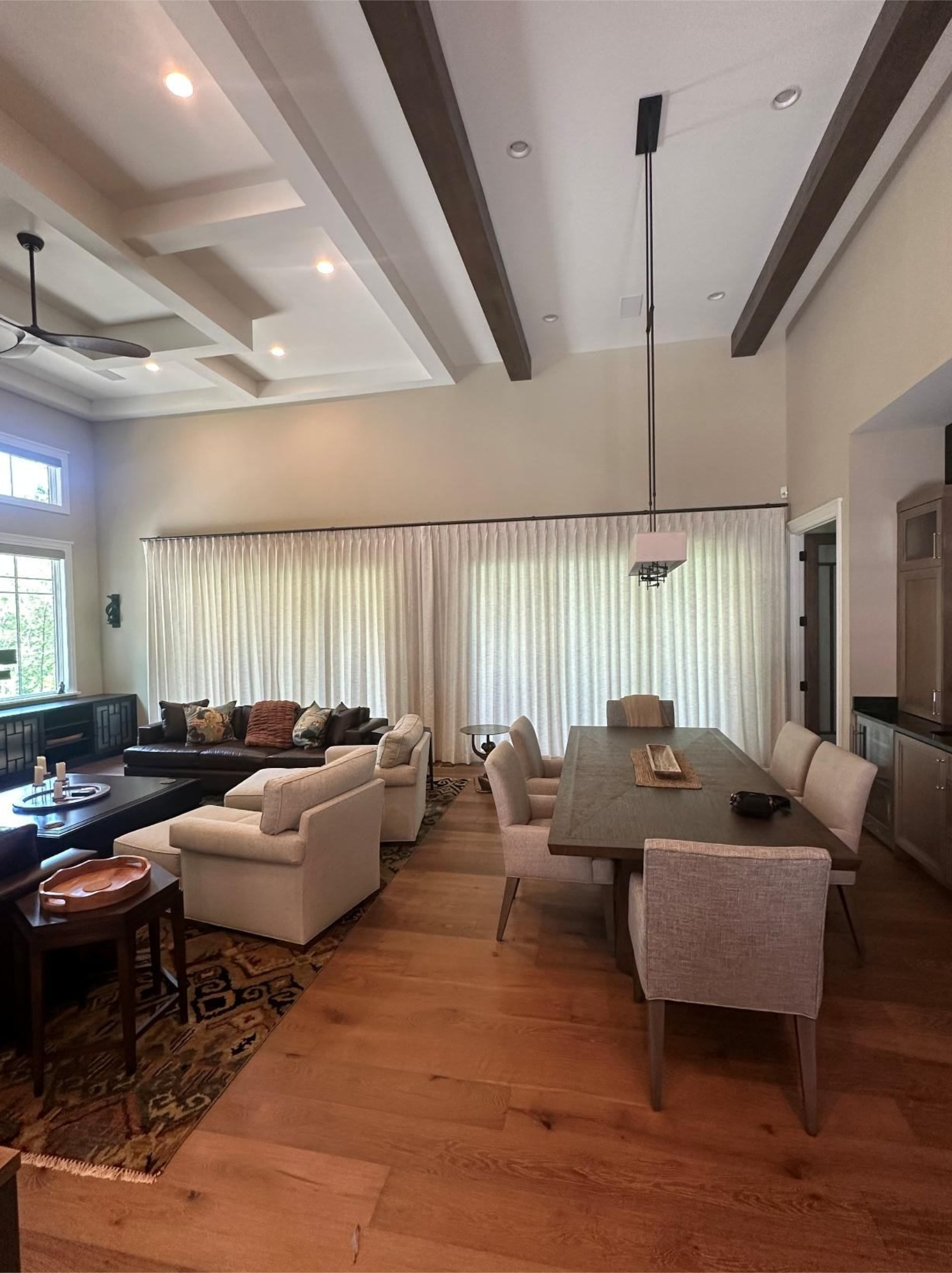 Bright living-dining room with beige sofas, a wooden table, high ceilings, and large curtained windows.