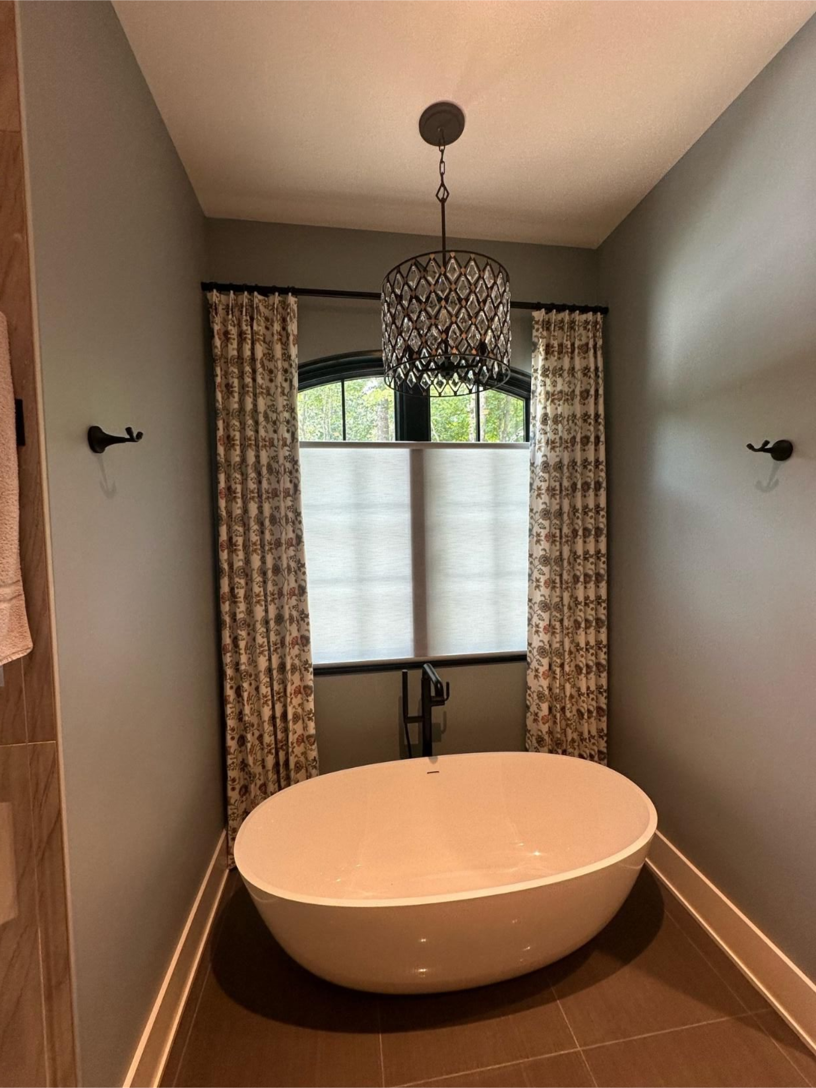 Narrow bathroom with a freestanding white tub, chandelier, and window with curtains and blinds.