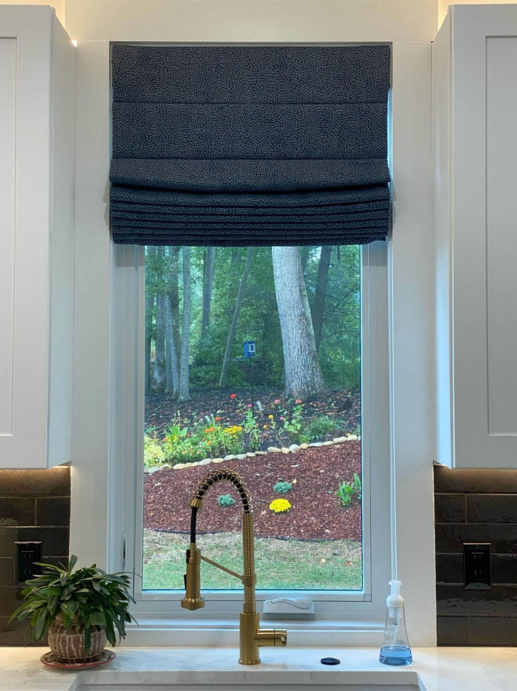 Kitchen window with dark Roman shade, brass faucet, and wooded backyard view.