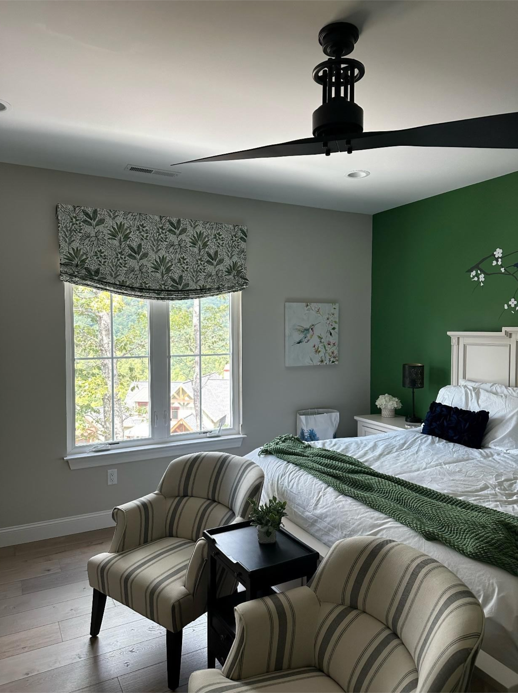 Bedroom with green accent wall, bed, window with patterned shade, and two striped chairs facing a small table