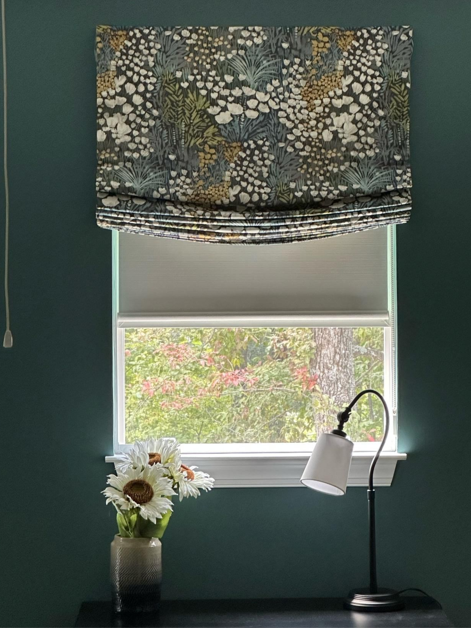 Window with gray blind, floral valance, vase of flowers, and desk lamp in a teal room.