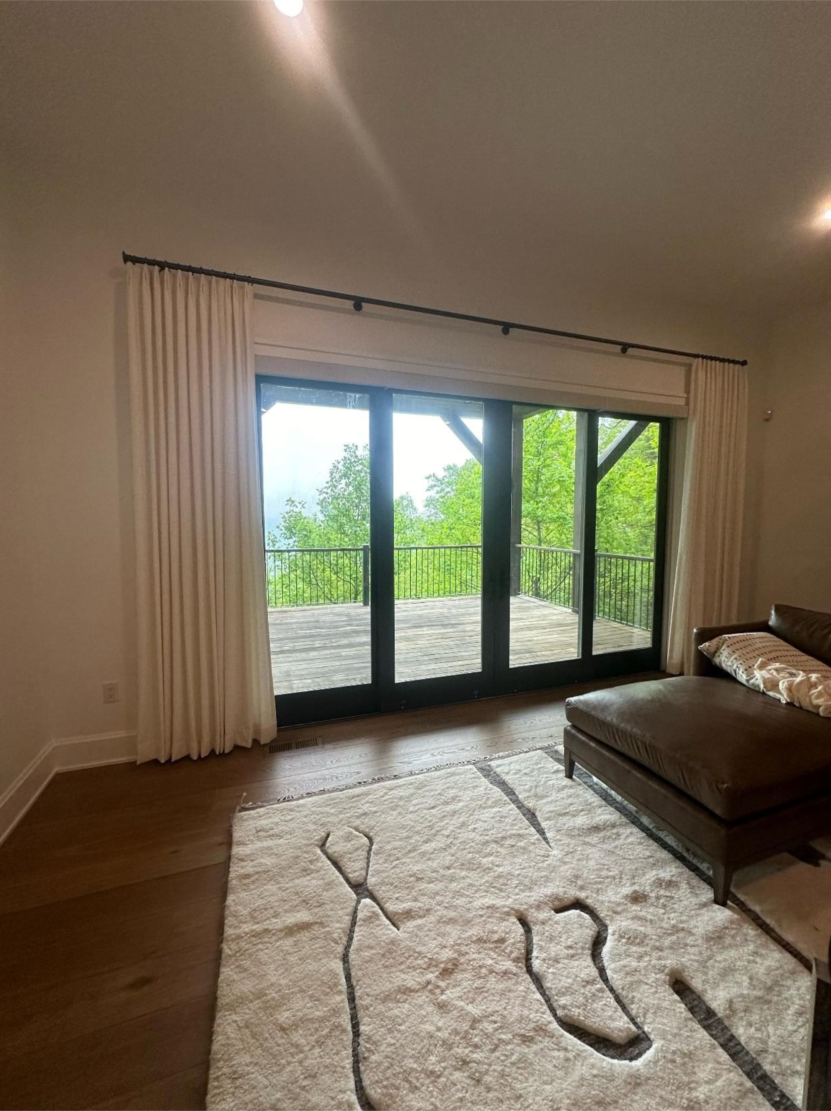 Bright living room with large glass doors, white curtains, beige rug, and dark leather seating.