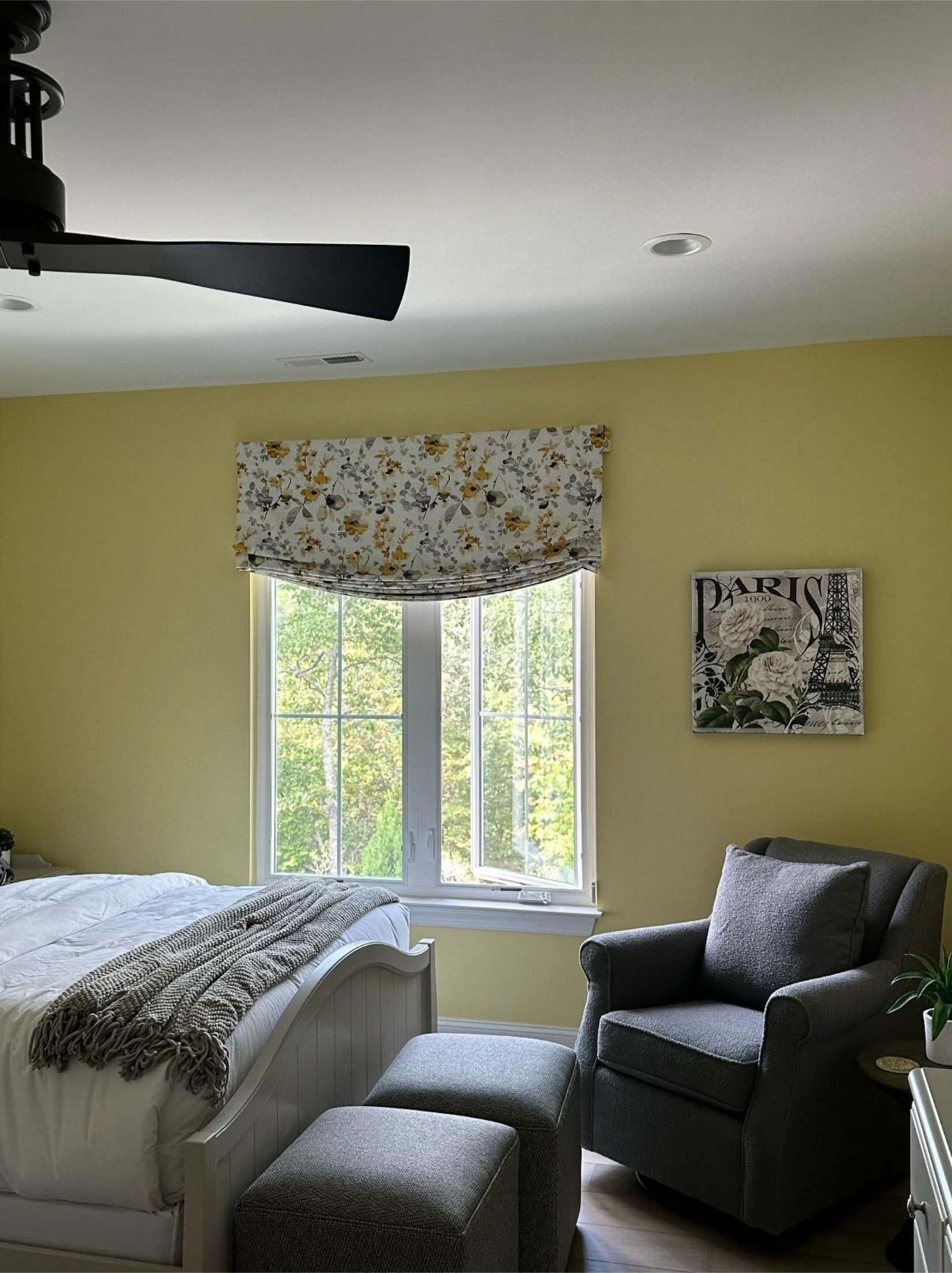 Bright bedroom with yellow walls, floral window valance, bed, and gray armchair by a sunlit window