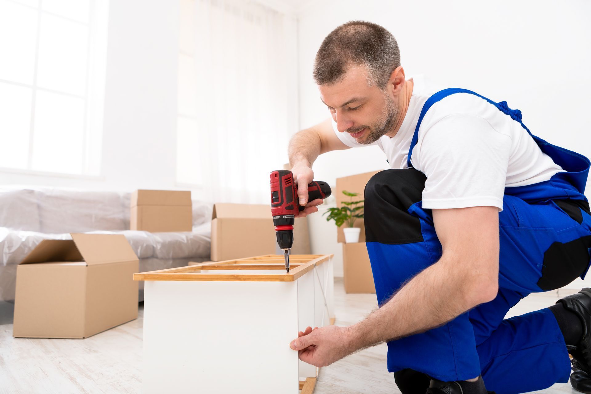 Man using a red drill to assemble a white cabinet in a bright room with cardboard boxes nearby