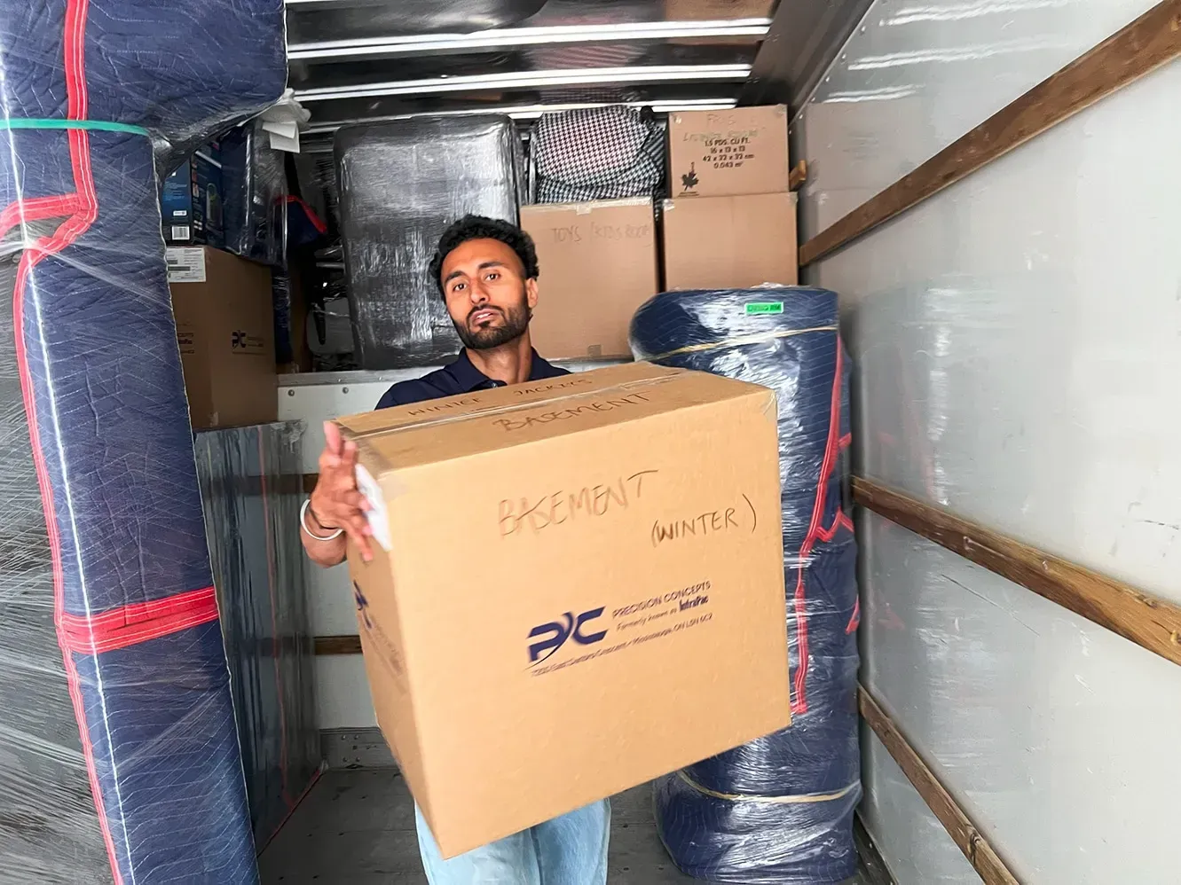 Man in a truck trailer carrying a large cardboard box among stacked wrapped pallets and boxes