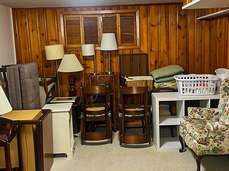 Cluttered room with stacked chairs, lamps, and a floral armchair against wood-paneled walls