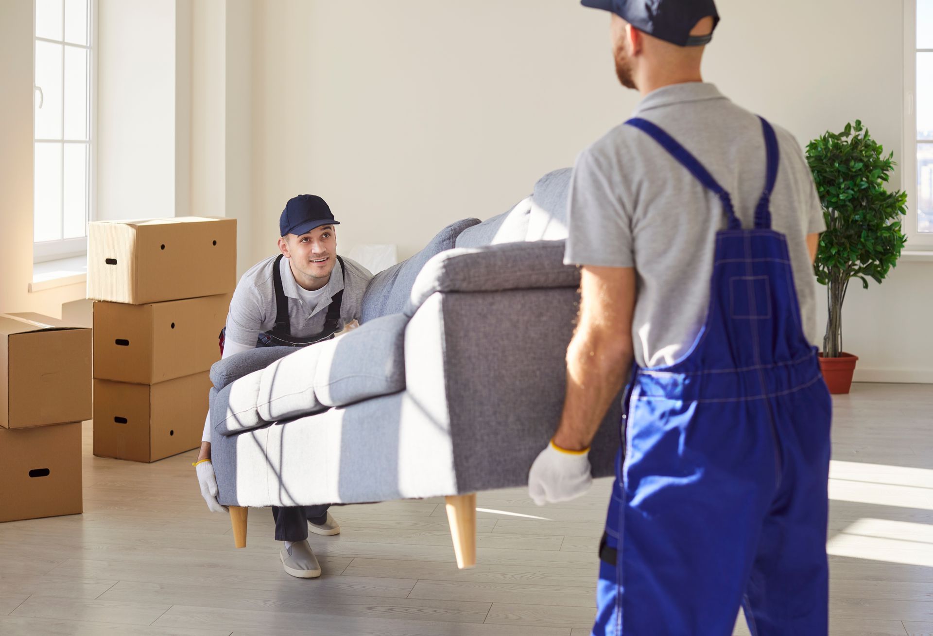 Two movers carrying a gray sofa through a bright room with moving boxes.