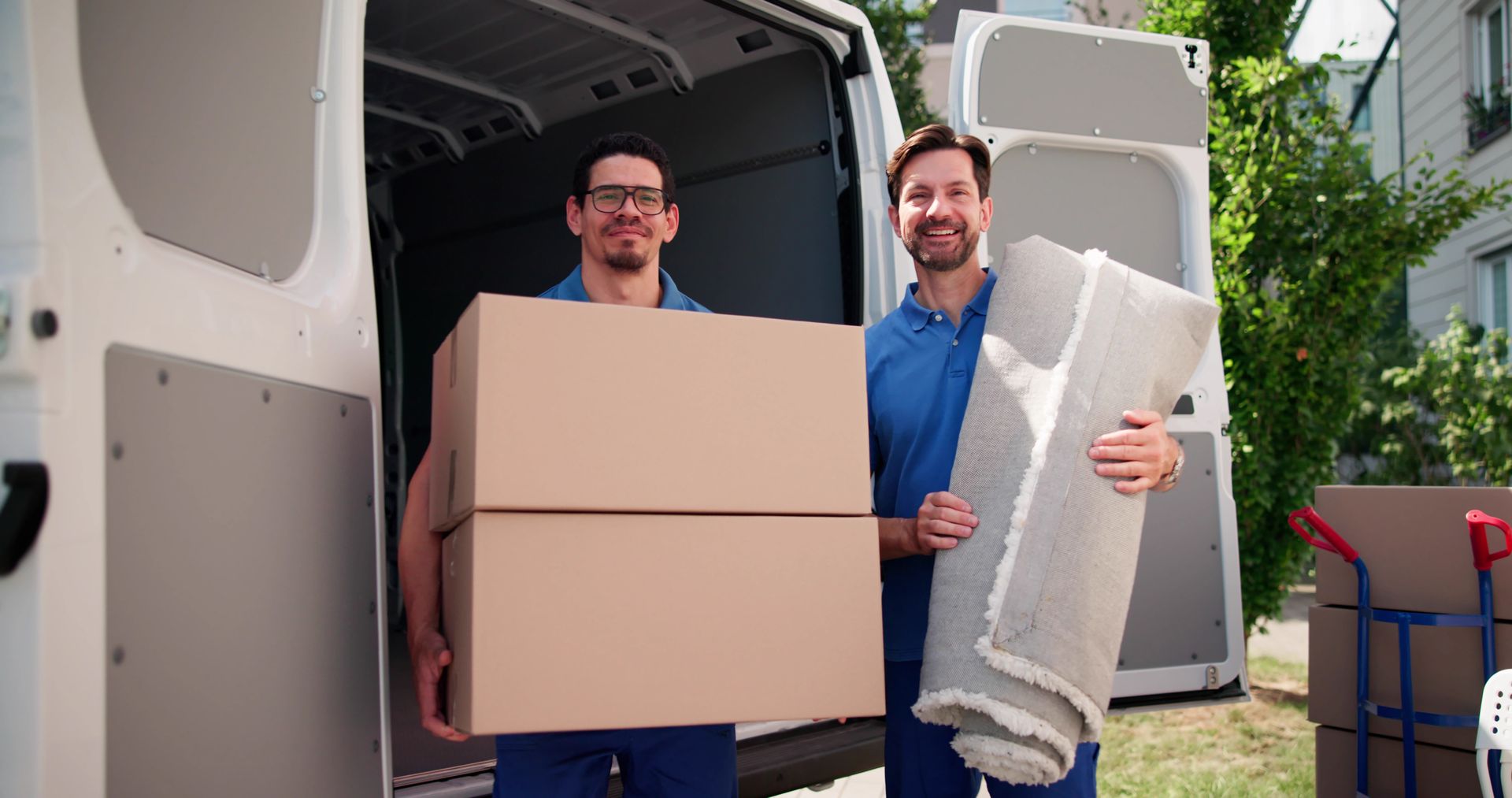 Two movers unloading cardboard boxes and a rolled rug from a white van outdoors.