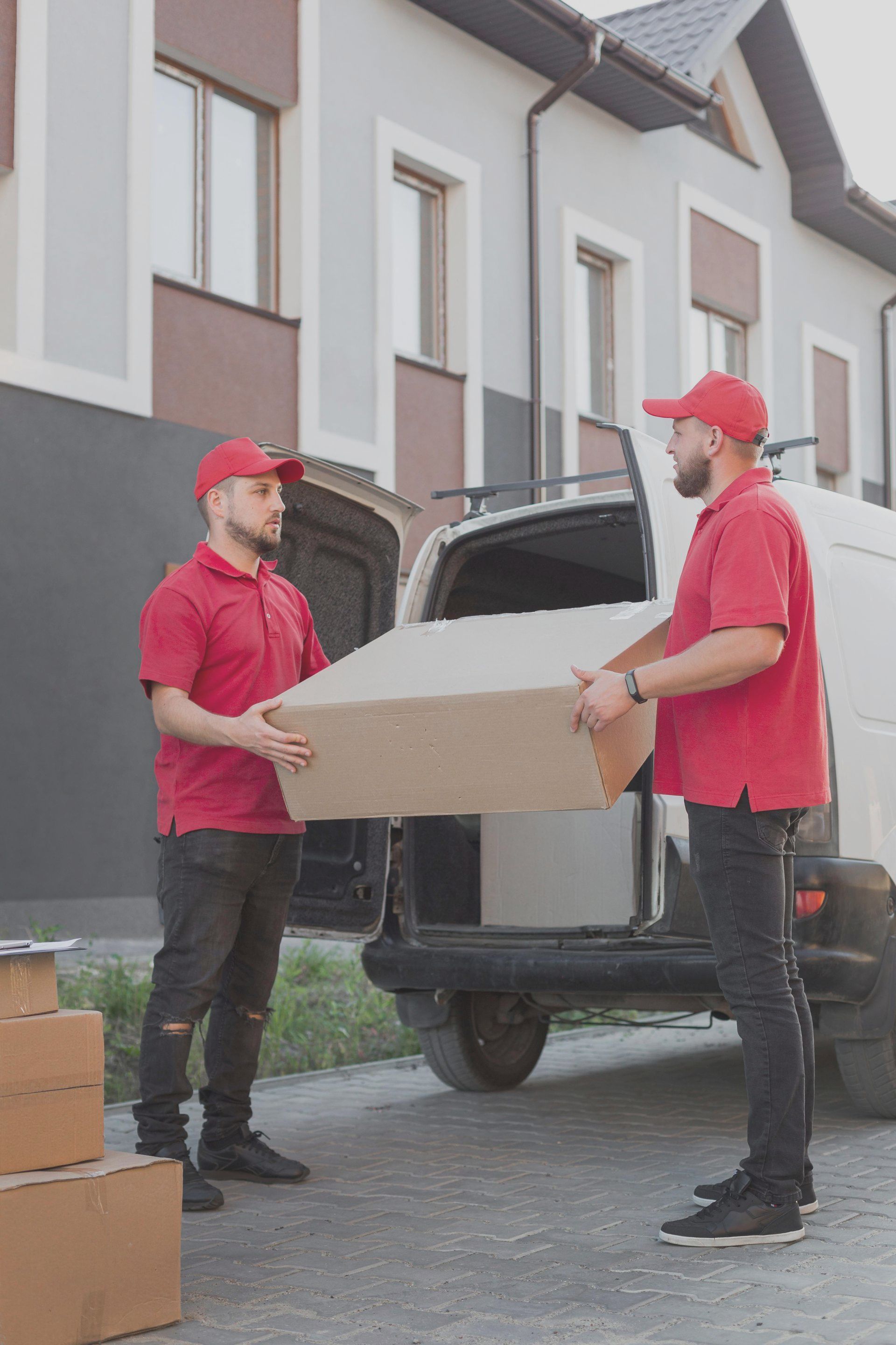 Two delivery workers in red uniforms carrying a cardboard box beside a van in a residential driveway