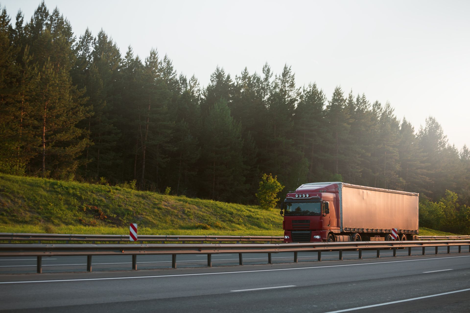Red semi-truck hauling a trailer on a highway beside a forested hillside at sunrise