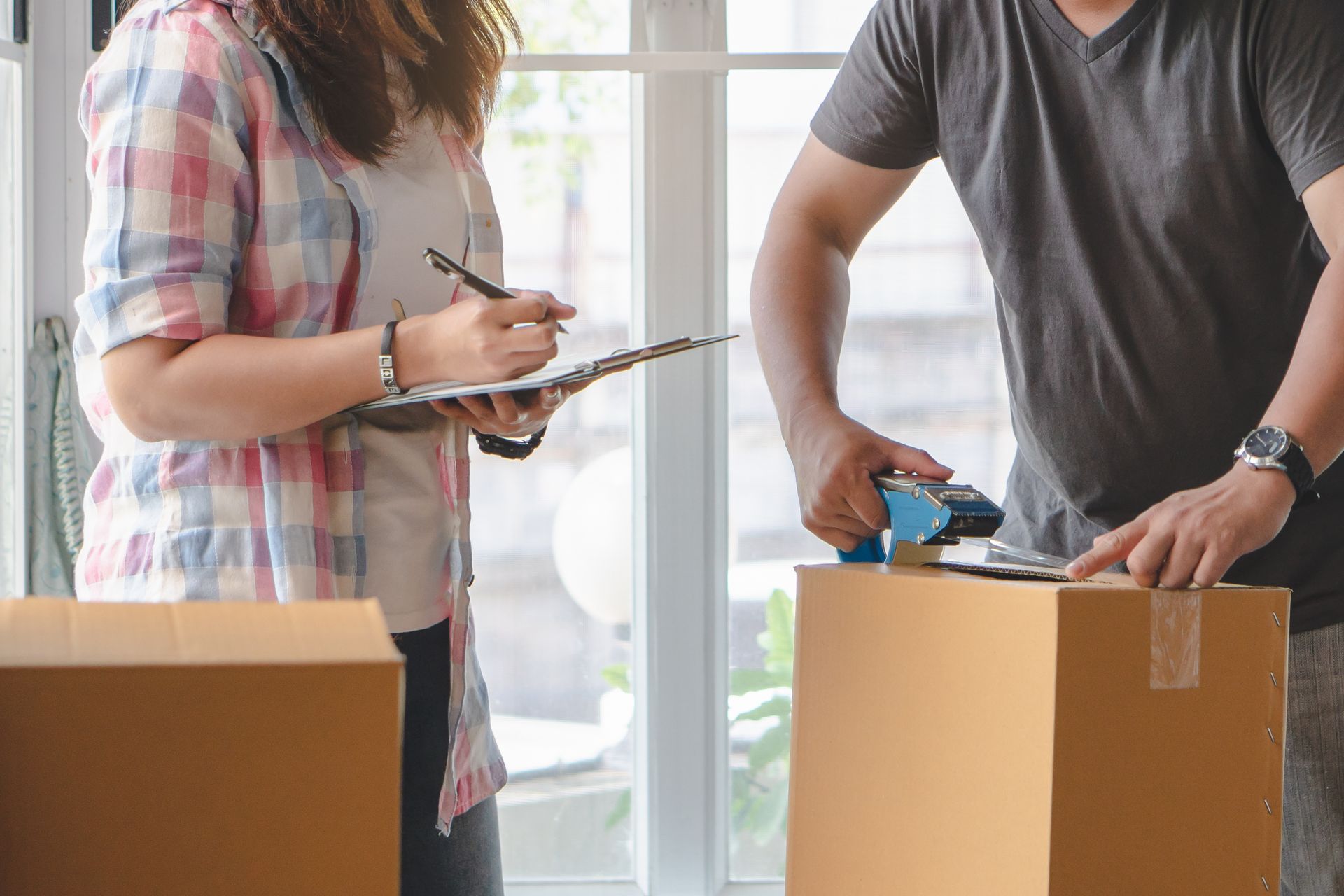 Two people packing a box with a clipboard and tape in a bright room