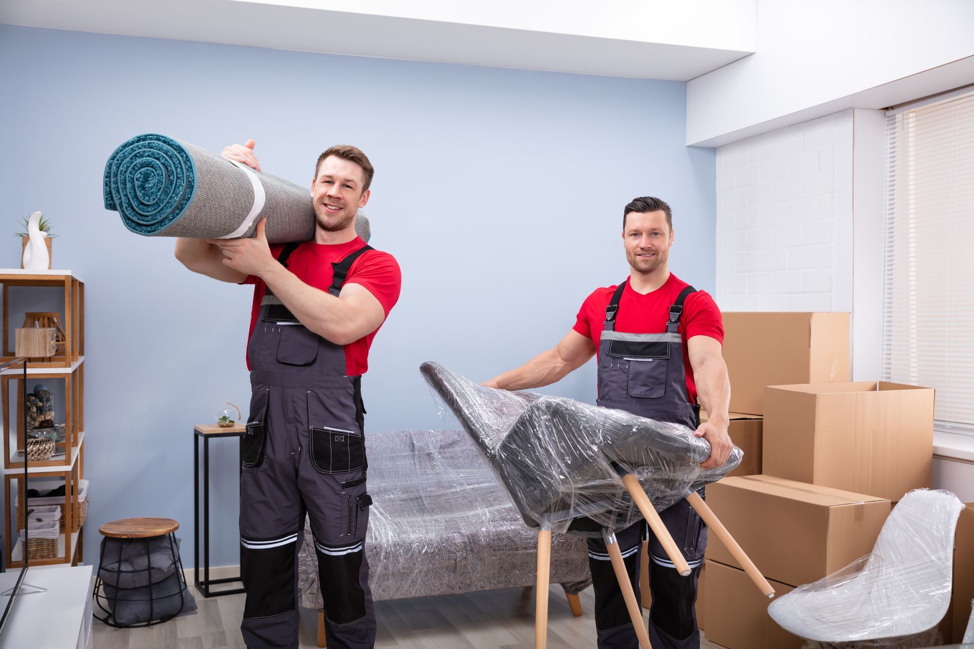 Two movers in red shirts carry wrapped furniture amid boxes in a bright room.