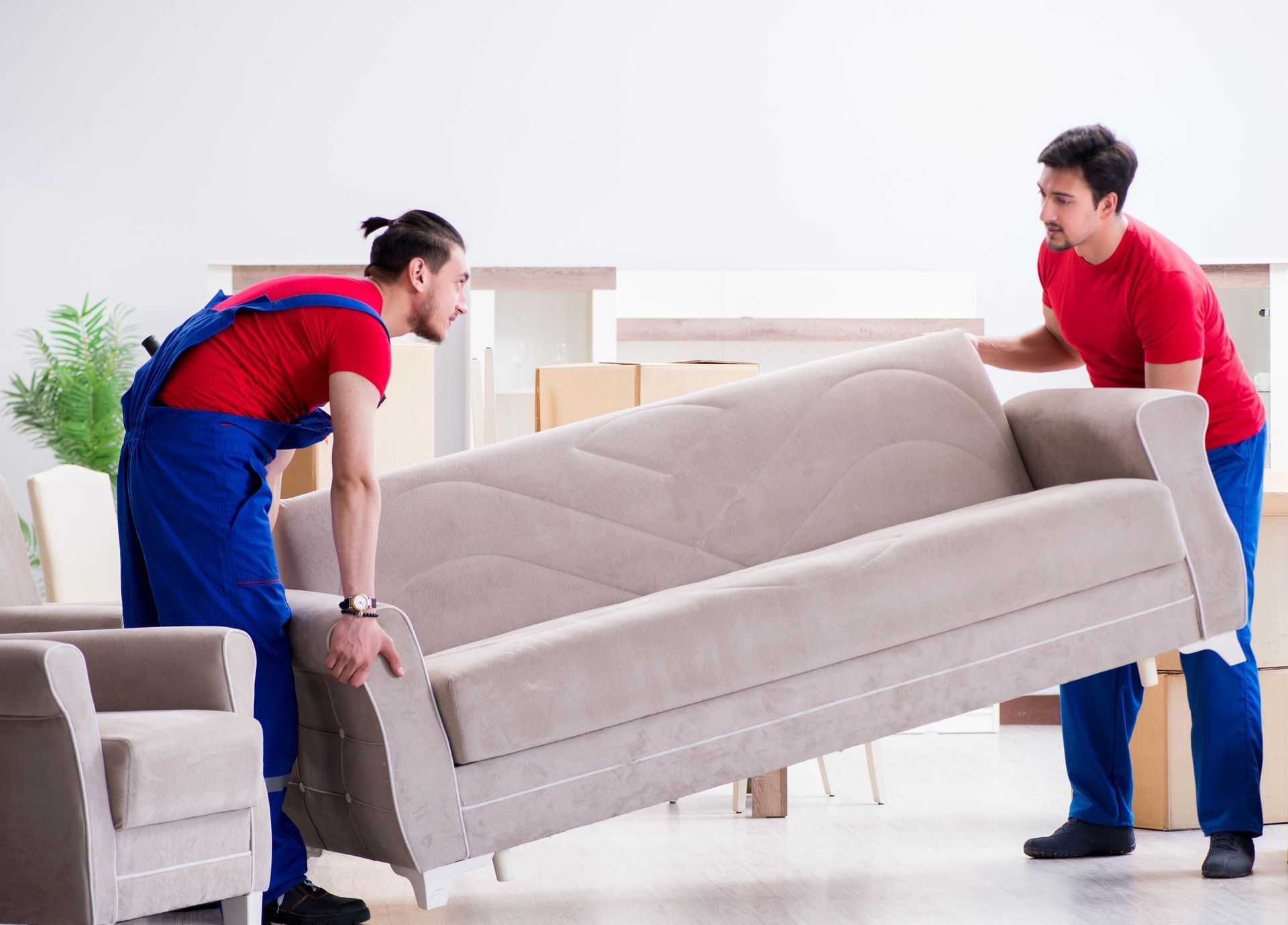 Two movers in red shirts and blue overalls carrying a beige sofa in a bright room