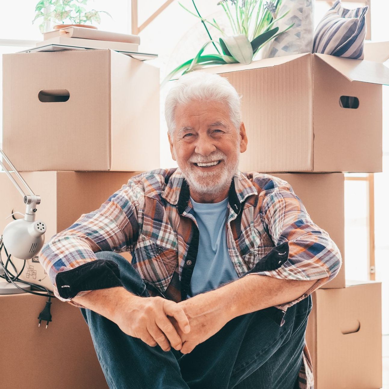 Smiling man sitting among moving boxes in a bright room, suggesting a home move.