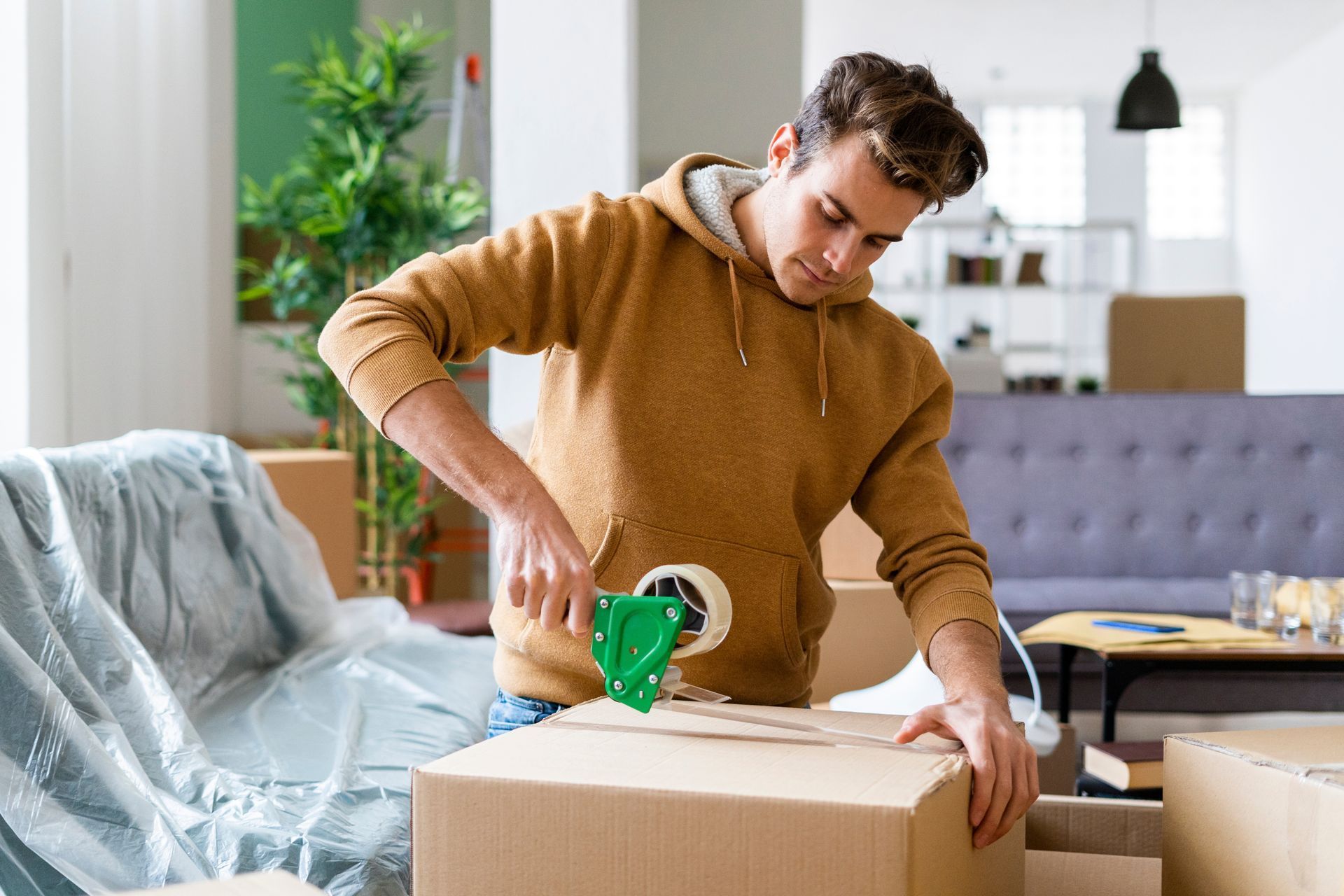 Man sealing a cardboard box with green tape in a bright living room